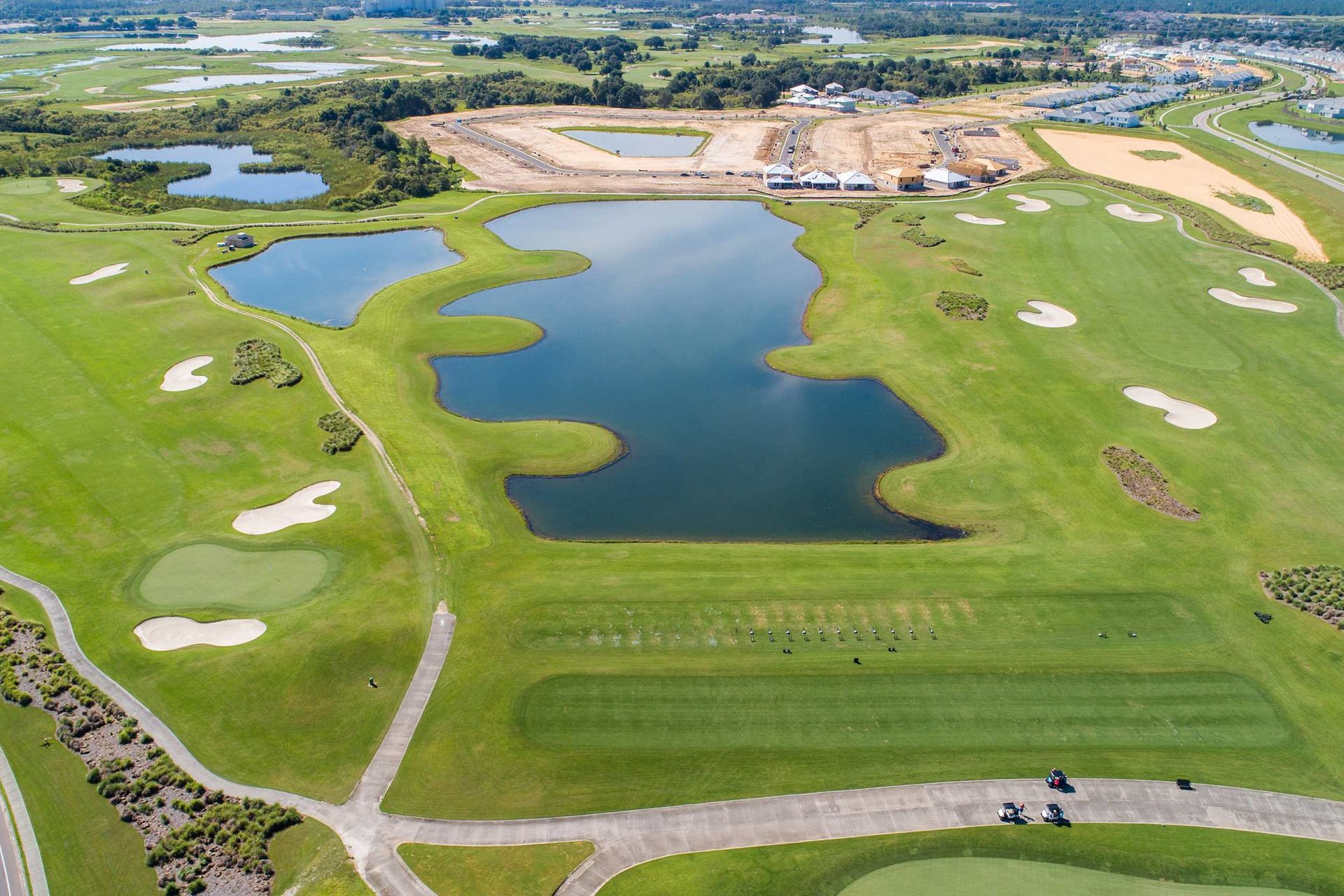 Aerial view of a sprawling golf course with lush greens, numerous sand bunkers, water hazards, and surrounding developments.