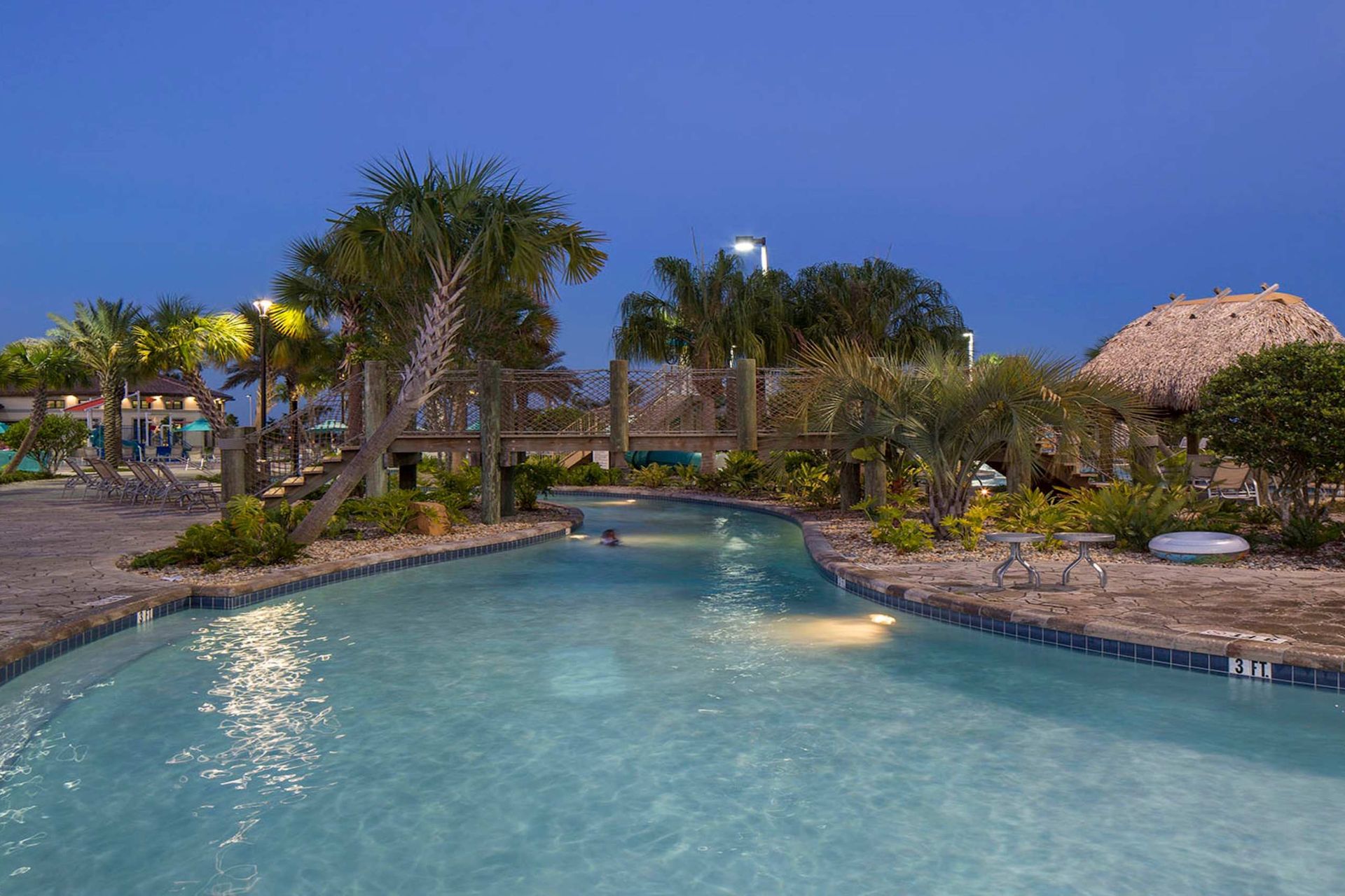 Resort-style lazy river illuminated at dusk, surrounded by tropical plants, palm trees, and a quaint wooden bridge.