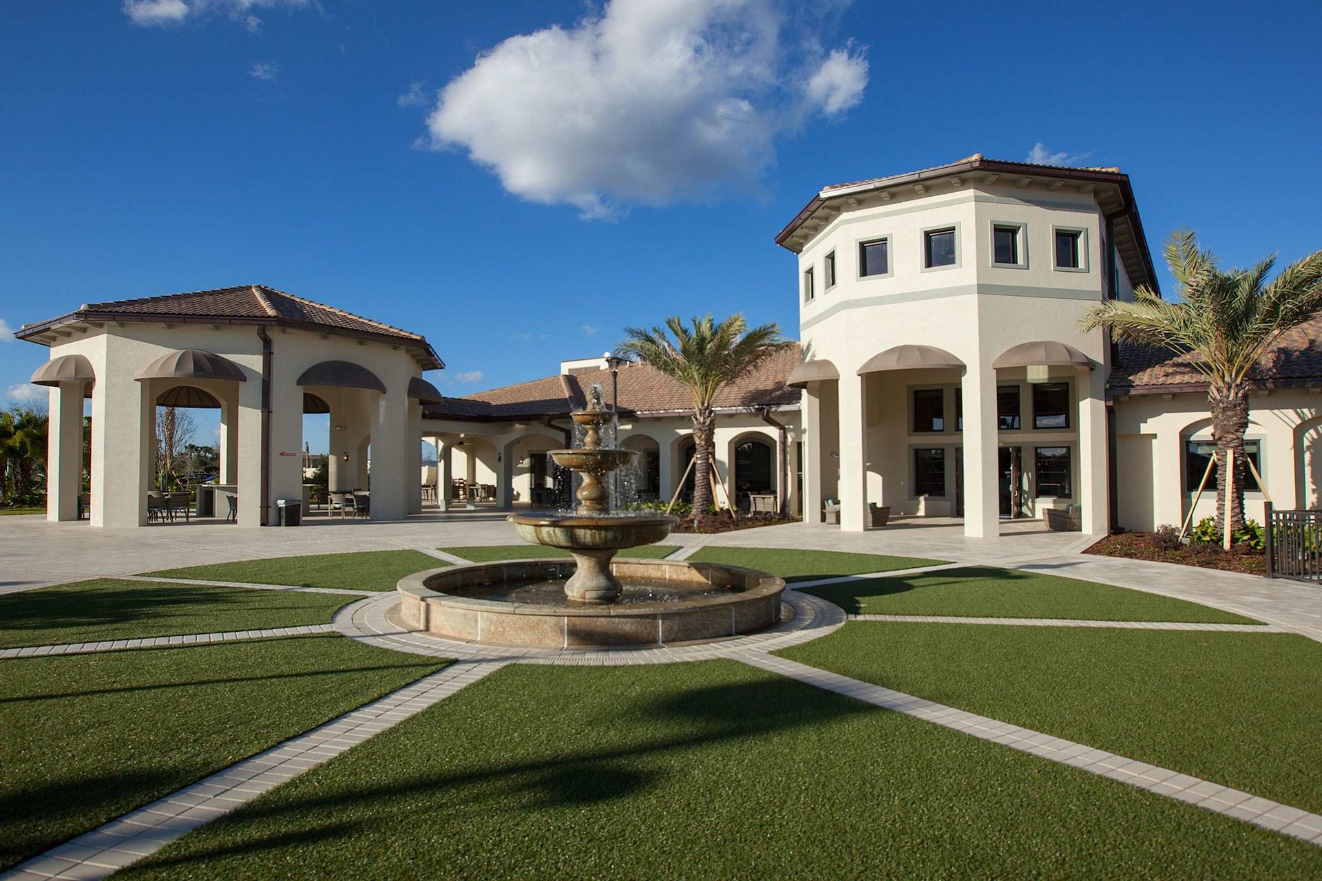 Elegant clubhouse exterior with a central fountain, palm trees, and arched walkways, under a bright blue sky.
