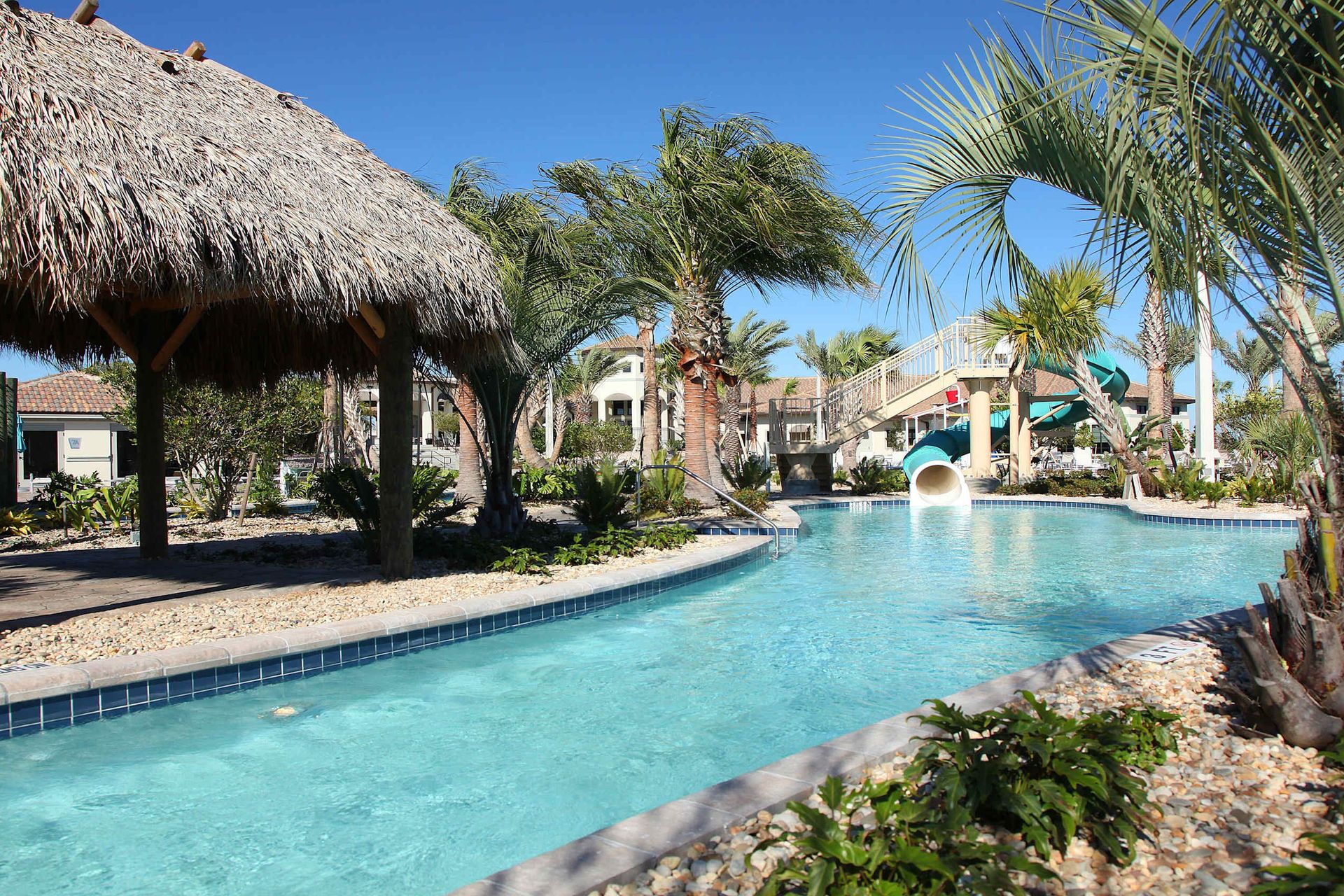 Resort-style pool with a lazy river, waterslide, lush tropical landscaping, and a thatched-roof cabana.