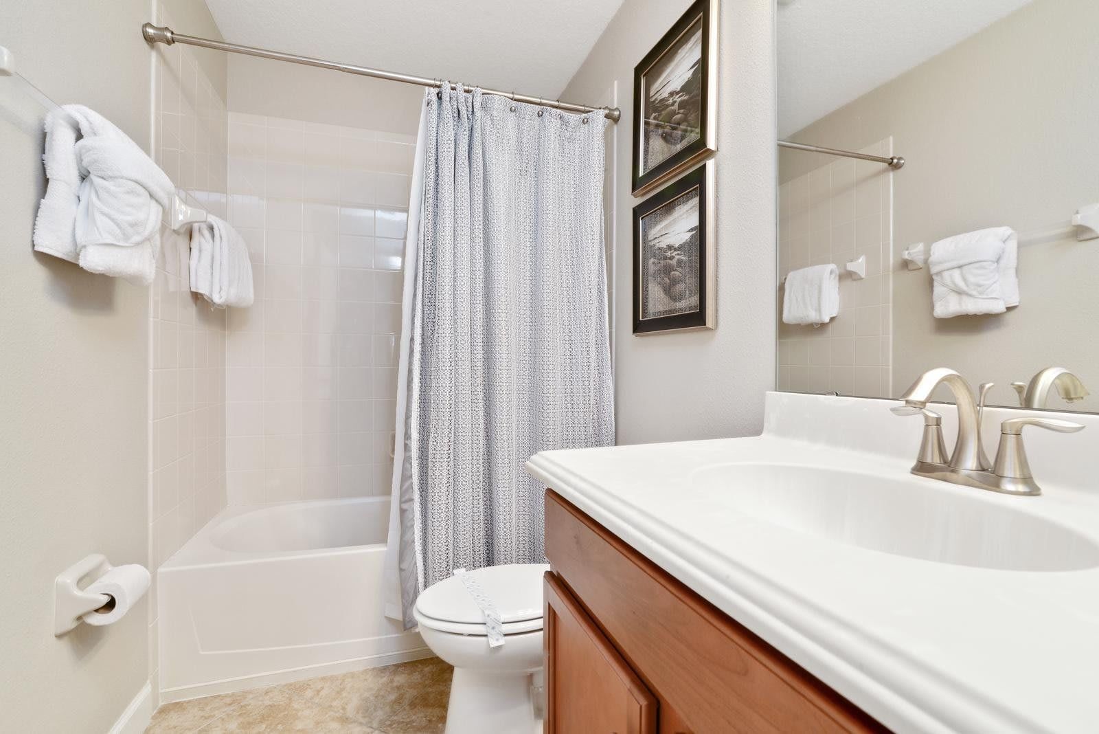 Cozy bathroom with a shower-tub combo, light tiles, soft white towels, and wood vanity under bright lighting.
