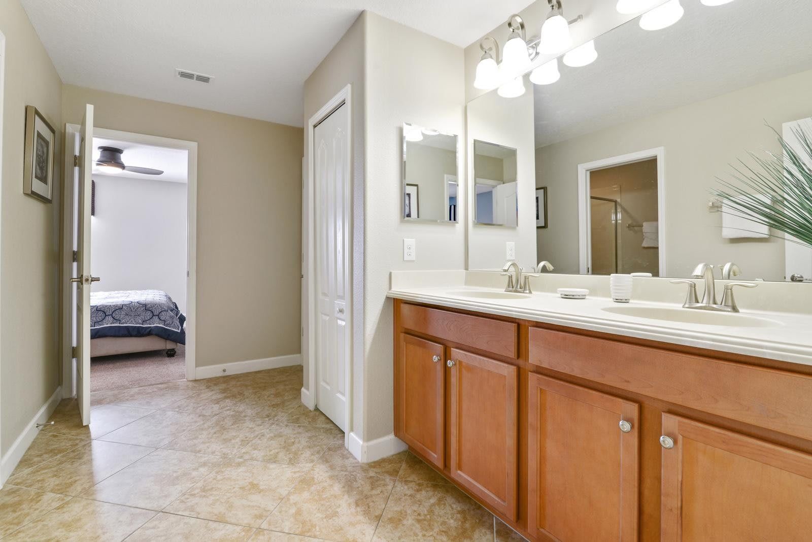 Spacious bathroom with double sinks, wood cabinets, bright lighting, and a tiled floor, leading to a bedroom.