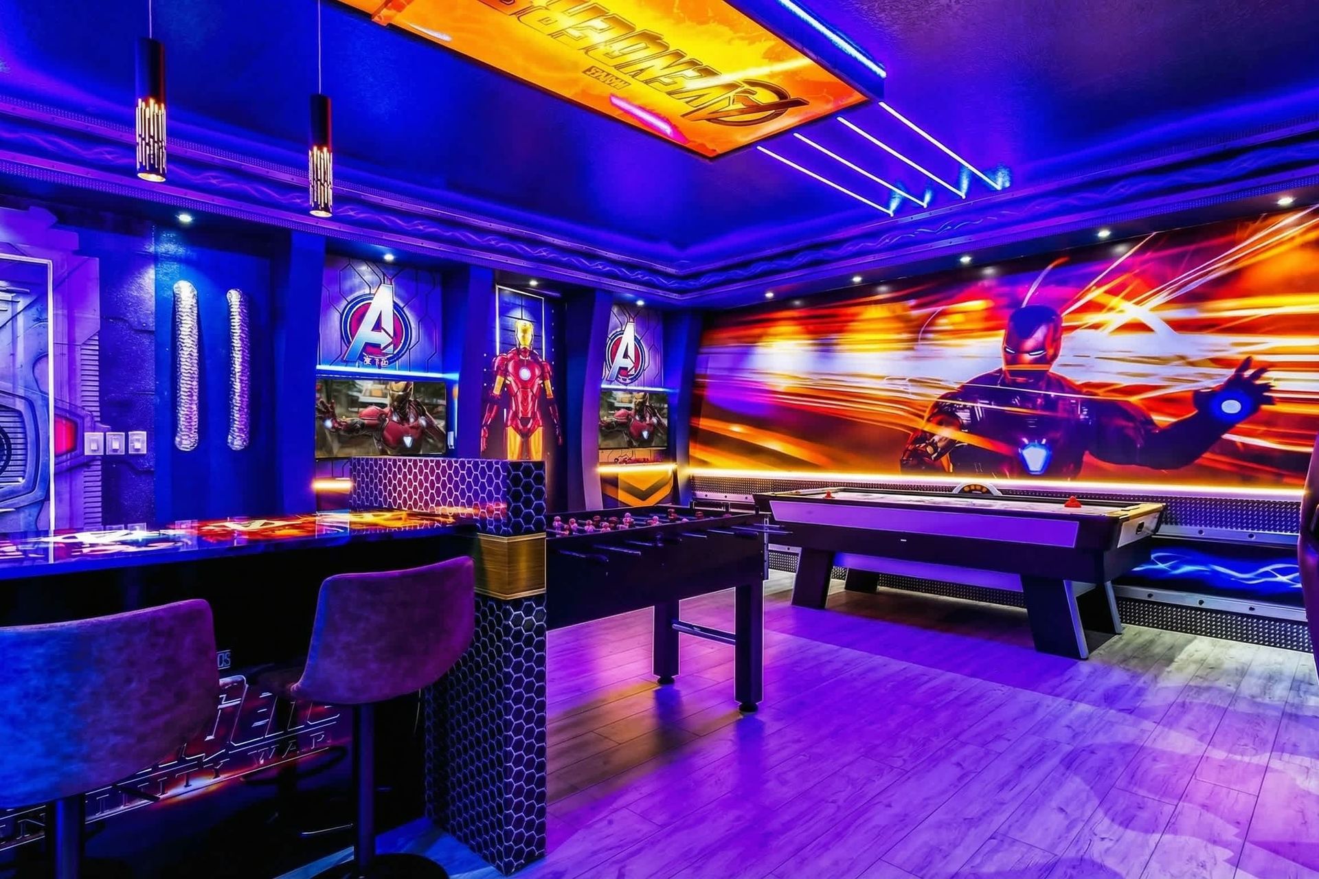 Avengers-themed game room with arcade games, air hockey, foosball, vibrant lighting, and superhero decor.