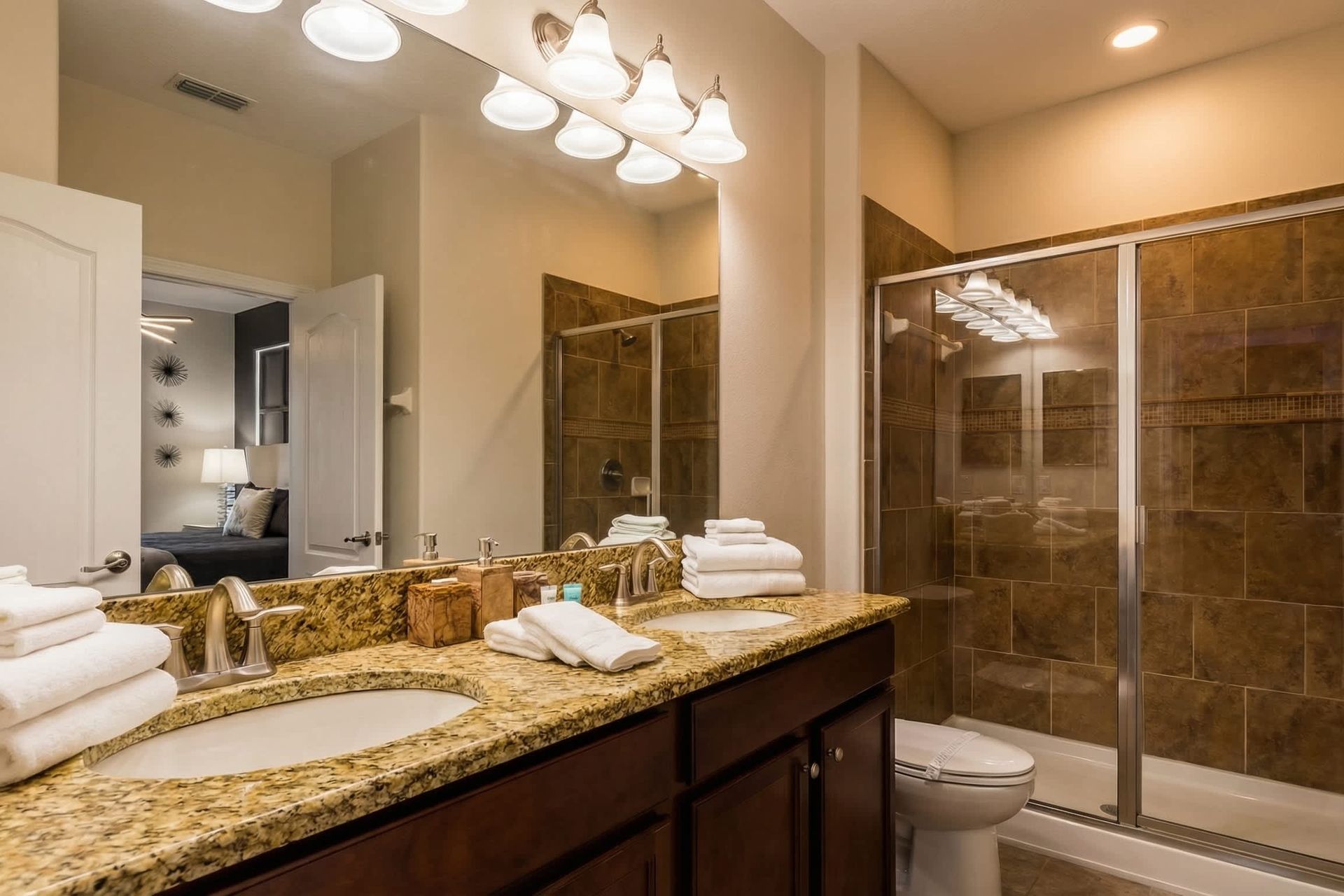 Modern bathroom with granite double sinks, wood cabinetry, tiled shower-tub combo, and elegant lighting.