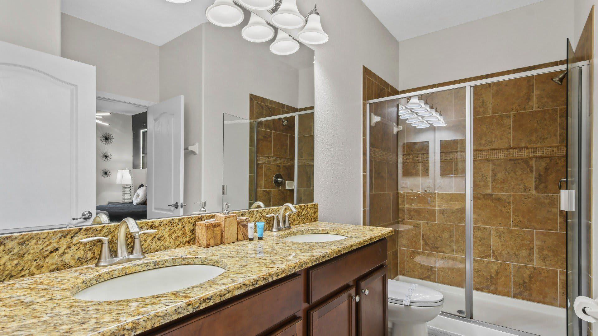 Modern bathroom with granite double sinks, wood cabinetry, tiled shower-tub combo, and elegant lighting.