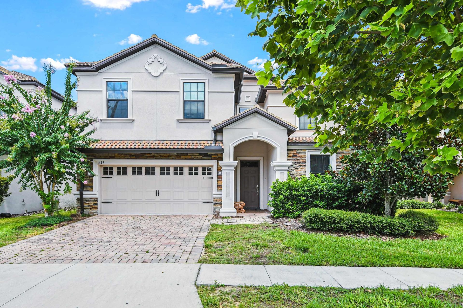 Beautiful two-story suburban home with a brick driveway, lush greenery, and a charming front porch.