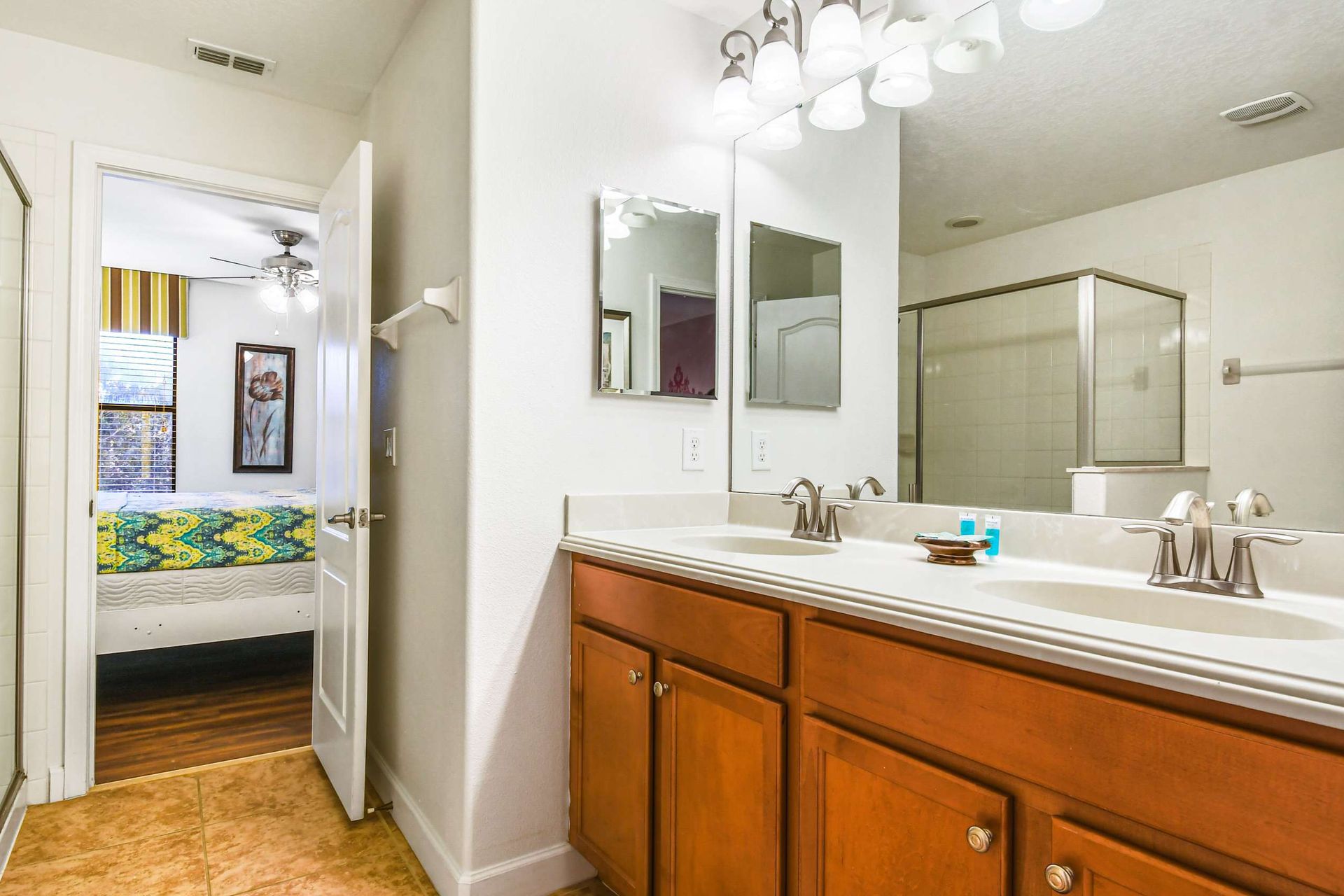 A bright bathroom featuring a double sink vanity, glass-enclosed shower, and a view into a cozy bedroom.