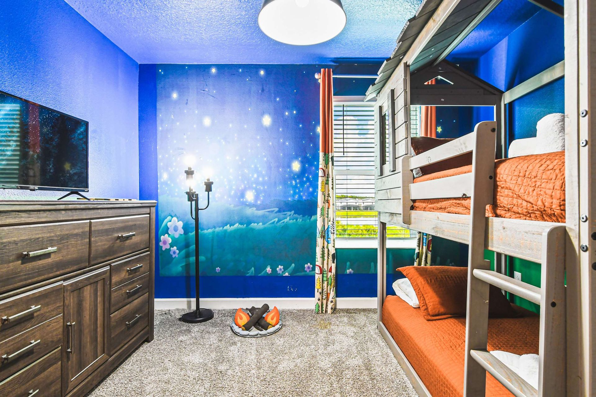 Whimsical kids’ bunk room with star mural, treehouse beds, campfire decor and TV
