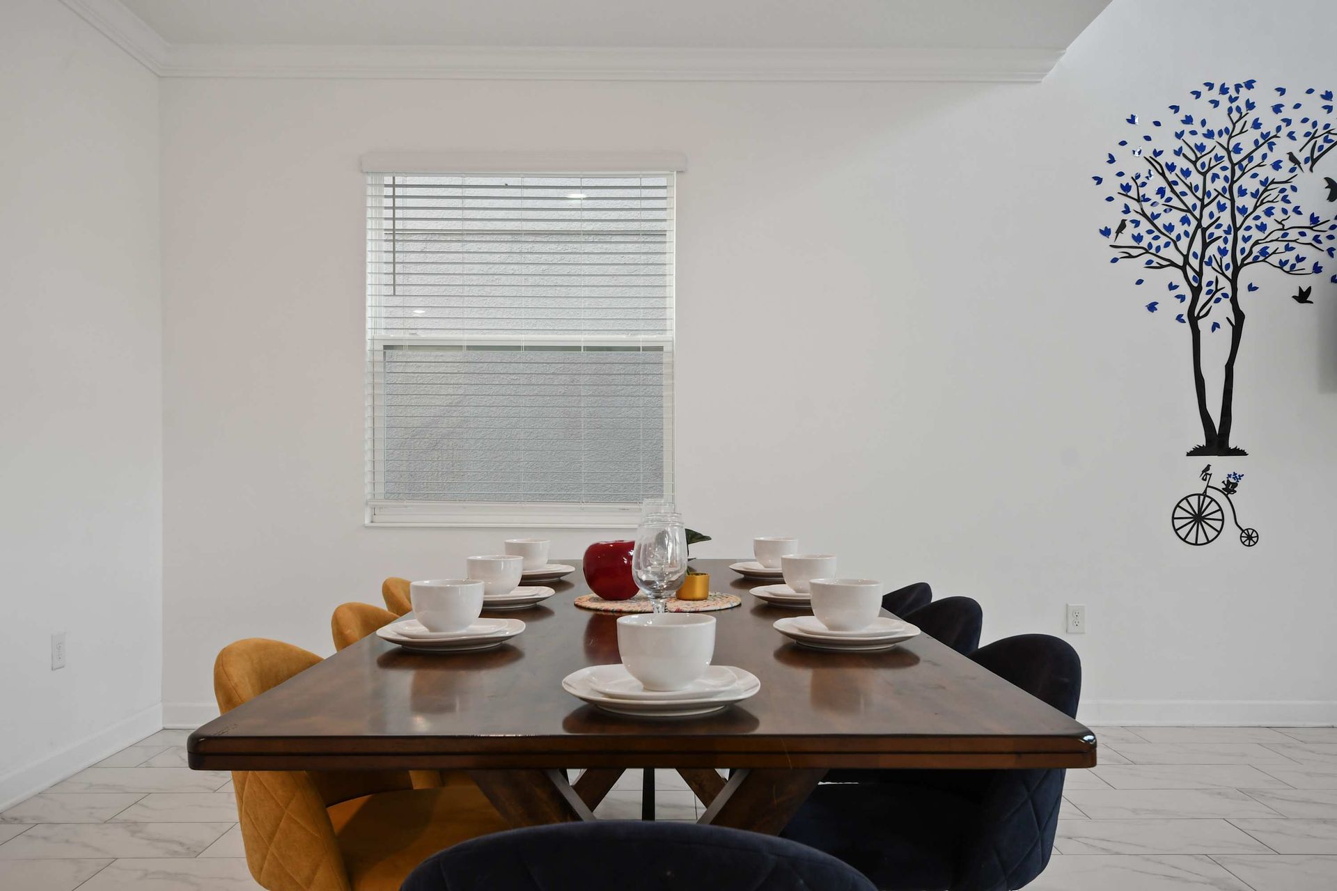 Modern dining table set for eight, with colorful chairs and whimsical wall art nearby.