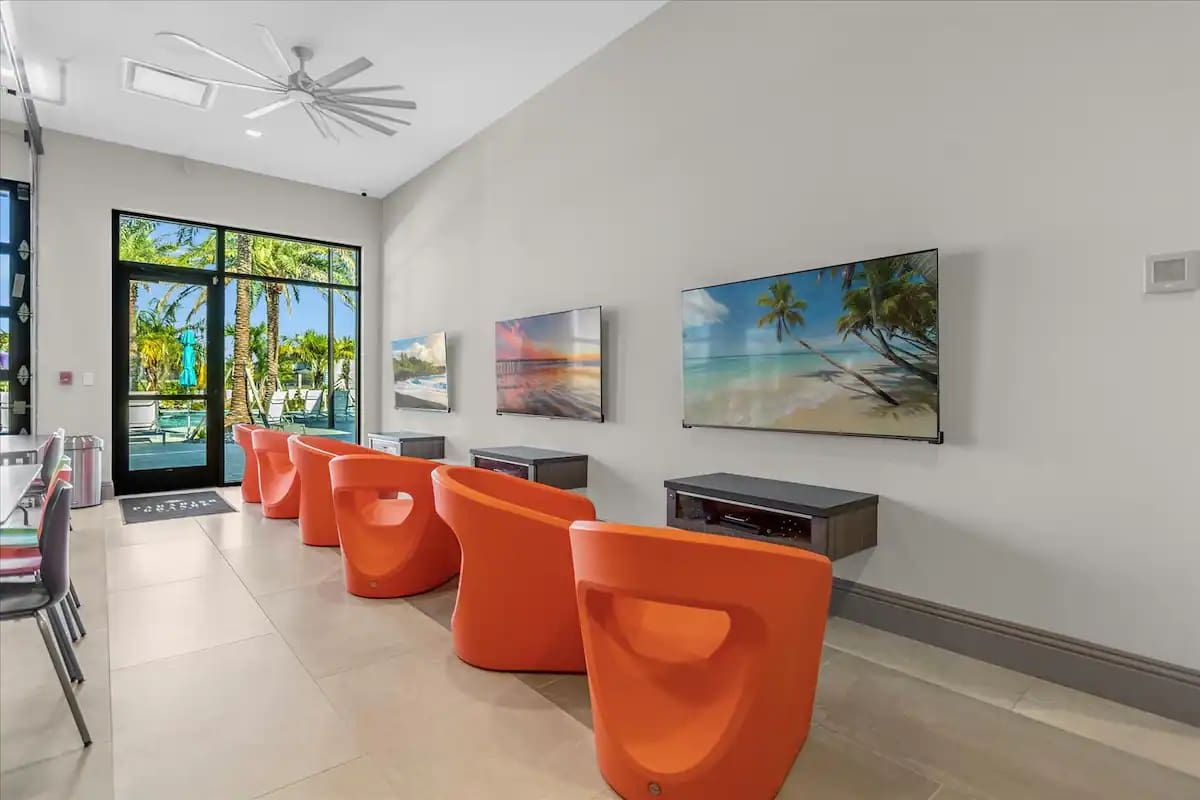 Colorful media lounge with gaming chairs, flat-screen TVs, and poolside views.
