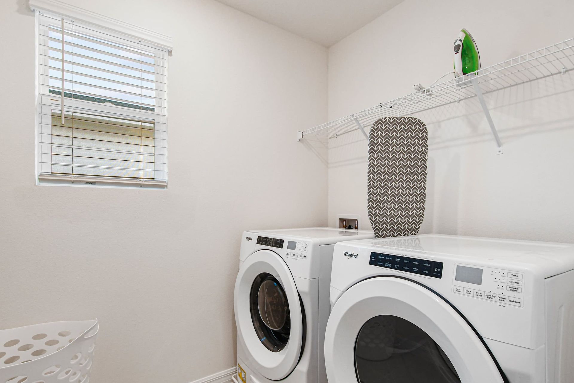 Bright laundry room with modern washer and dryer, ironing board, and storage shelf.