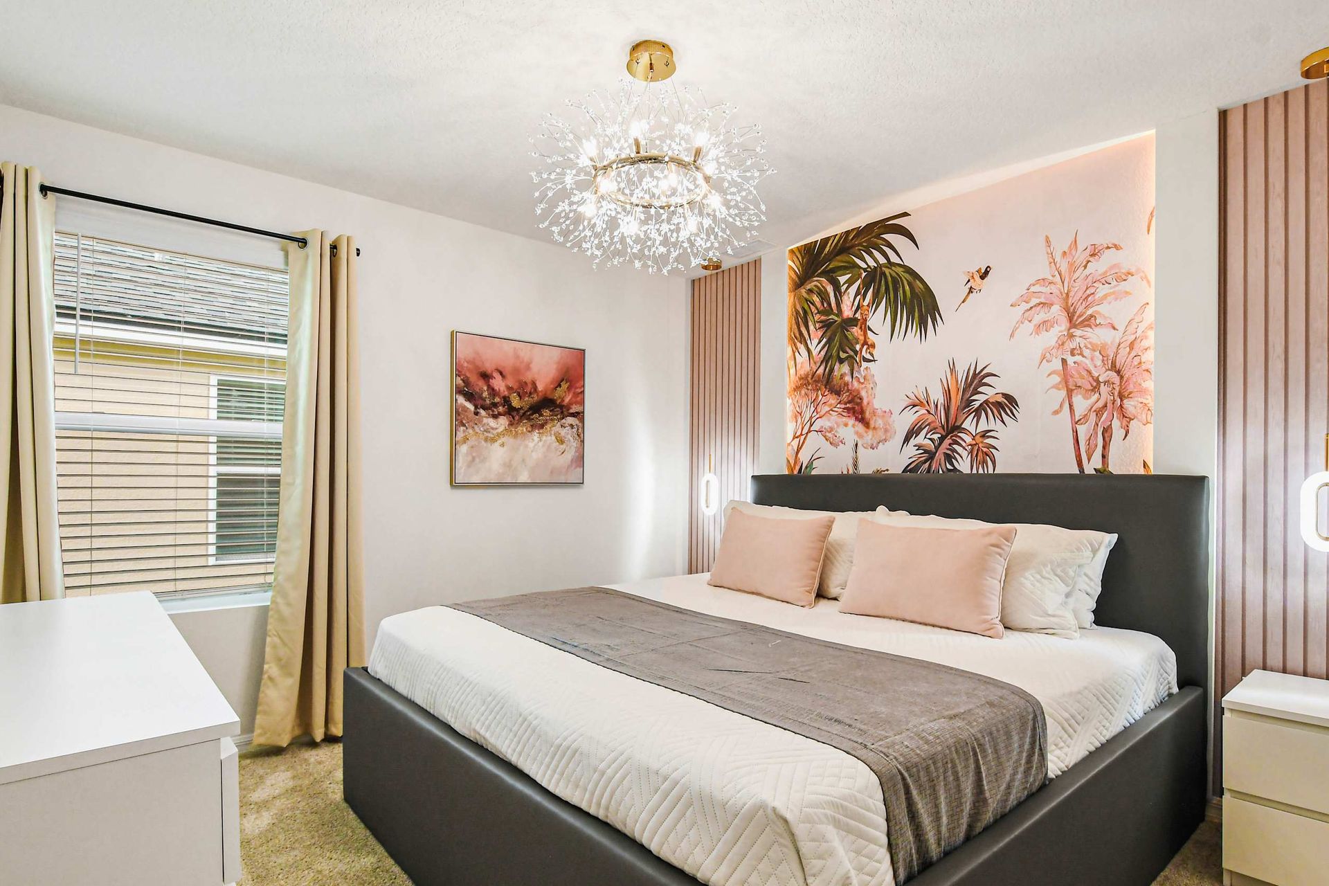 Elegant bedroom with blush accents, tropical mural, chandelier, and cozy king bed.