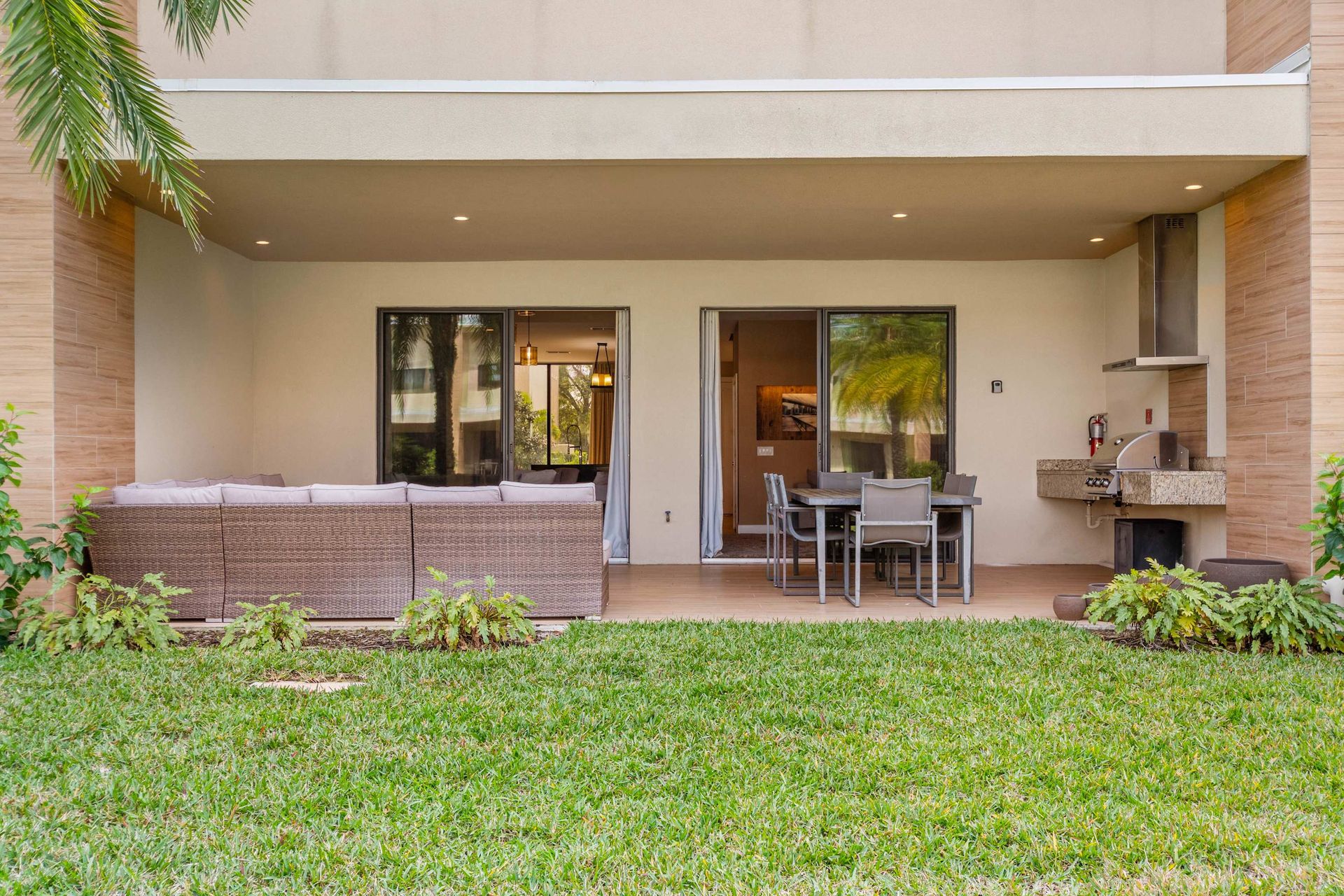 Covered patio with lounge seating, dining table, built-in grill, and lush backyard view
