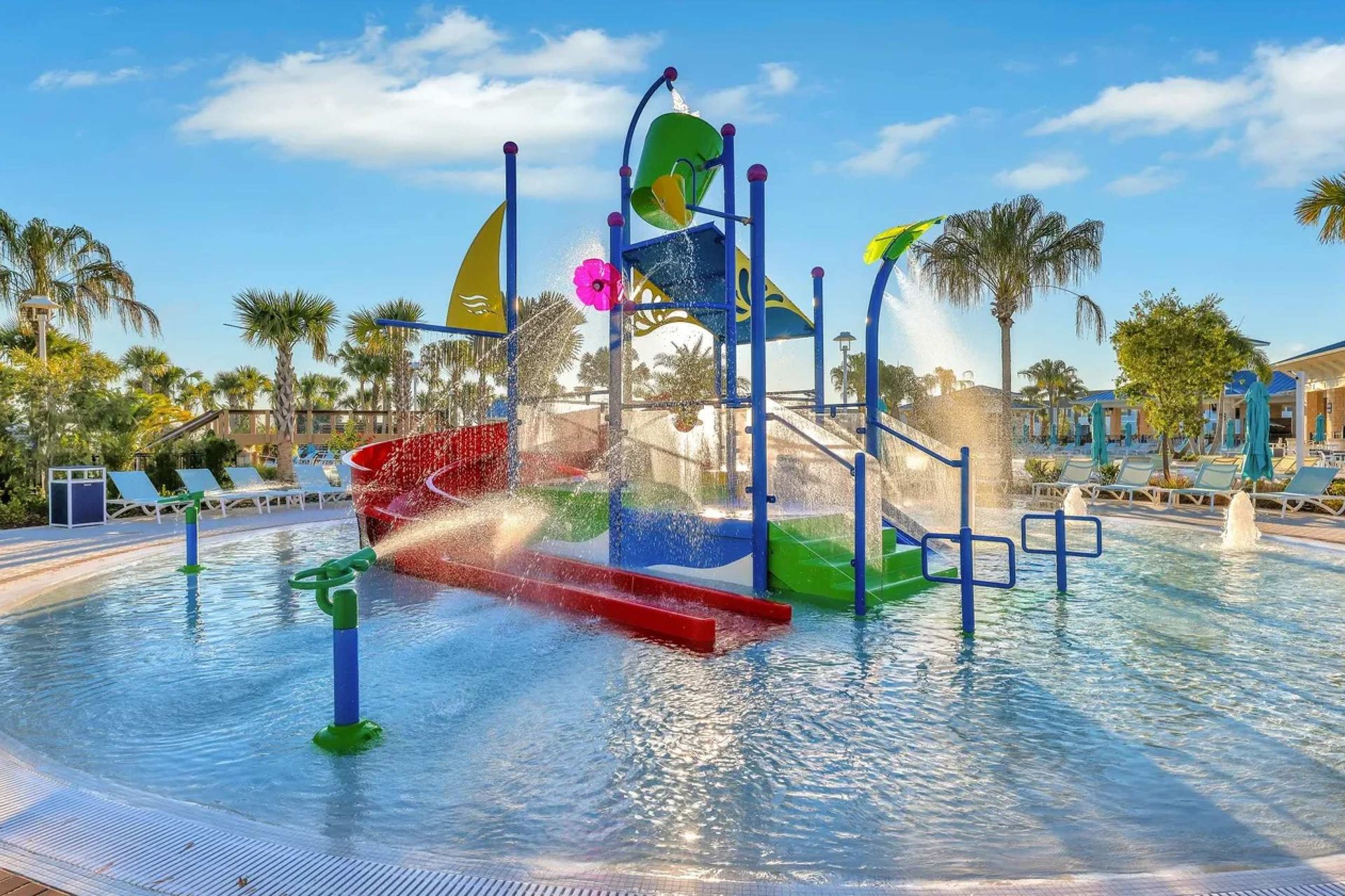 Colorful kids splash pad with slides, fountains, shaded cabanas, and lounge chairs
