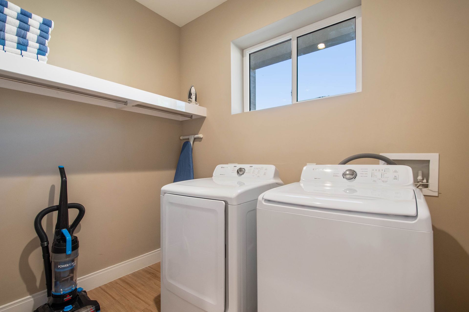 Bright laundry room with washer, dryer, storage shelf, vacuum, and natural light.
