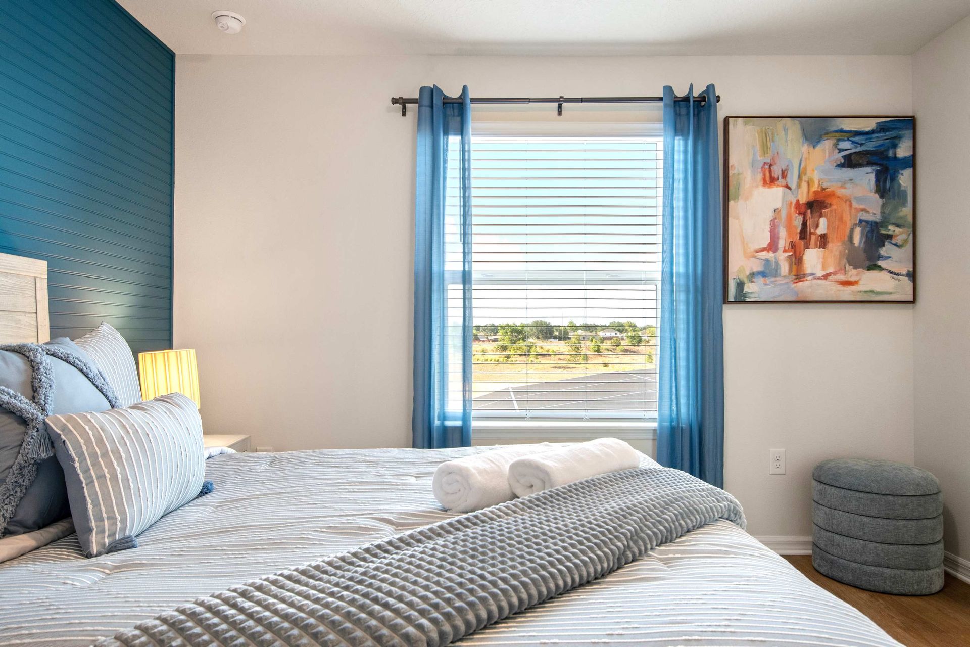 Sunlit guest room with cozy queen bed, artistic décor, and vibrant blue accents.
