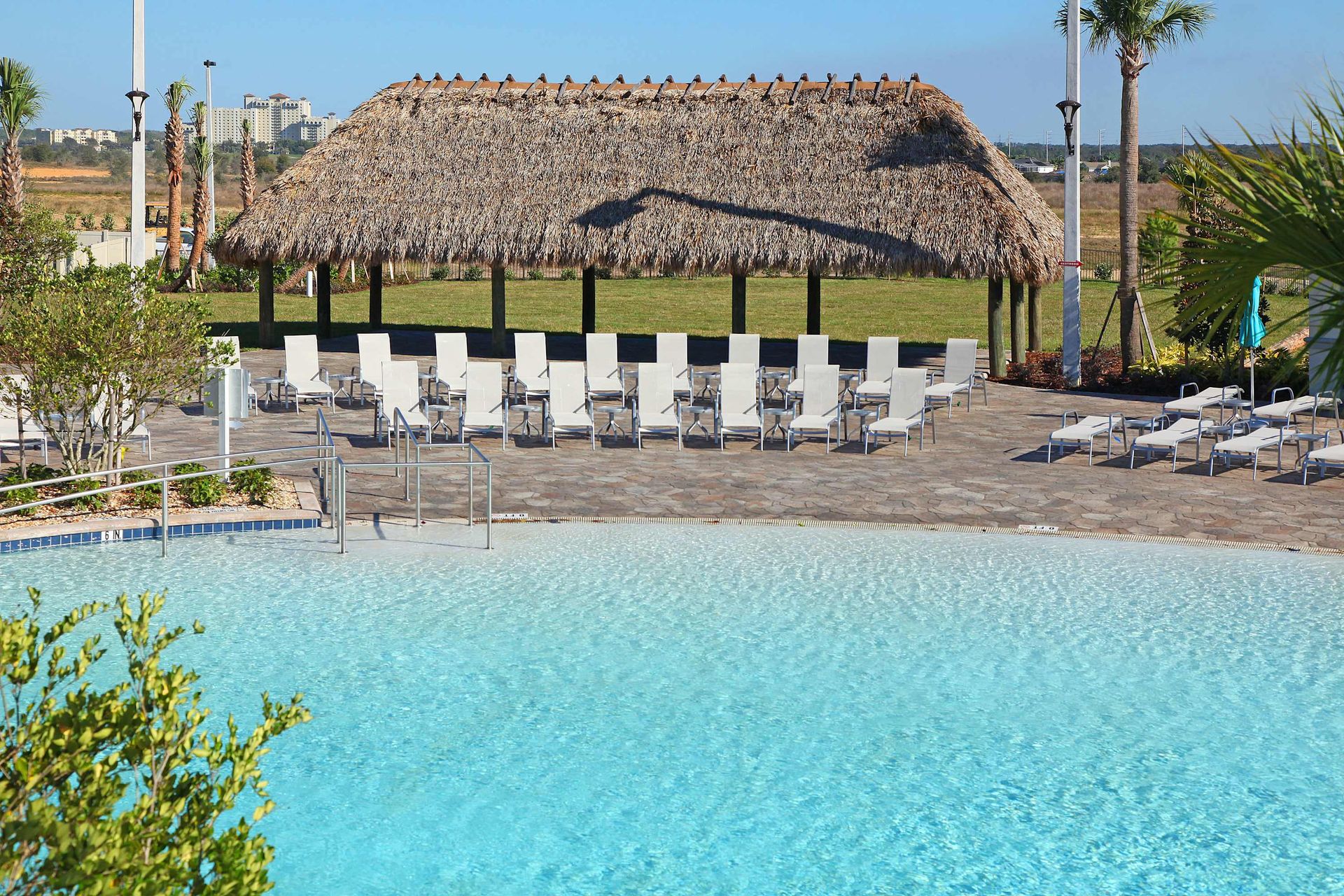 Bright sunny day at a tropical pool with a thatched roof and white furniture.