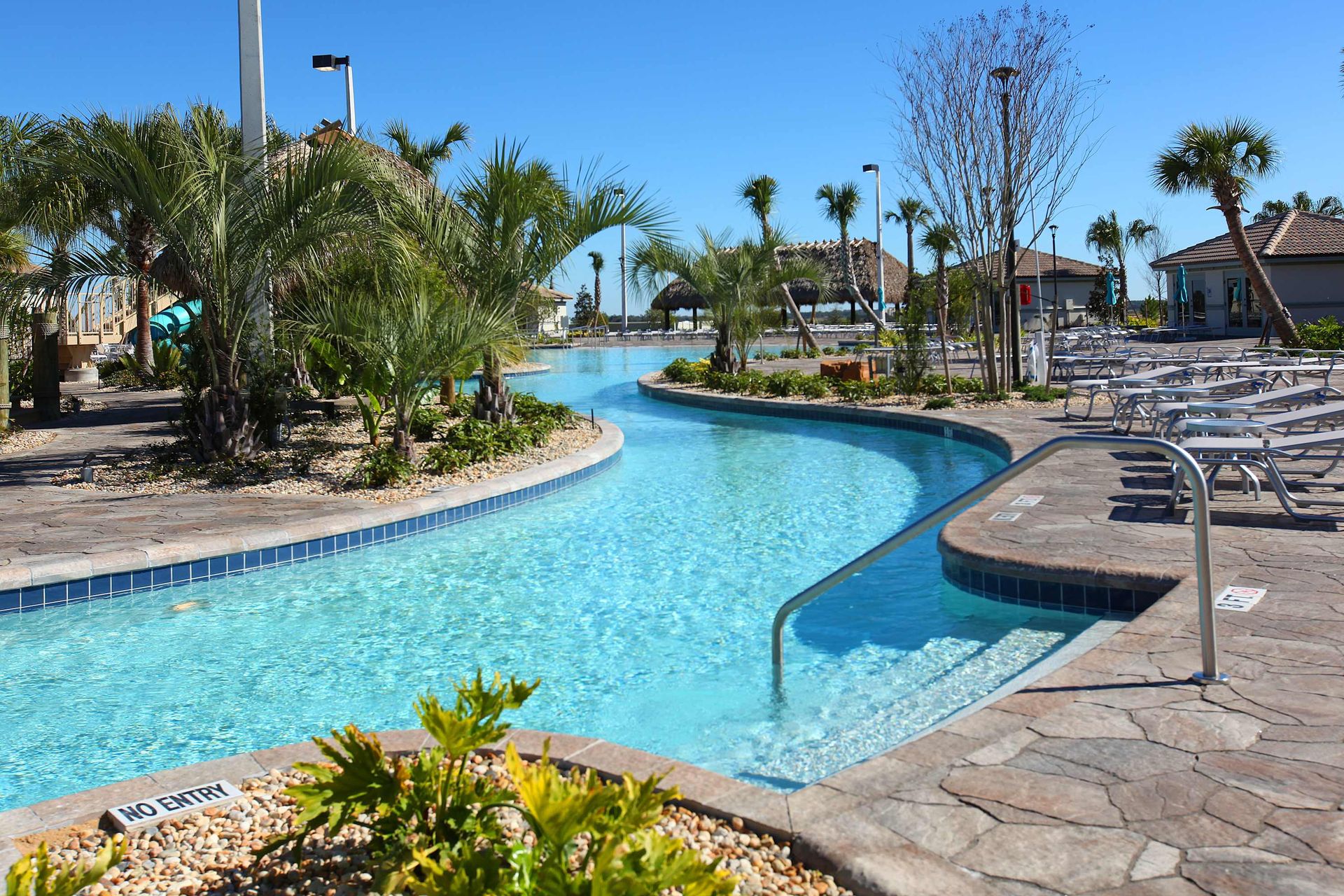 Sunny lazy river at a resort pool with clear water, handrails, and palm trees.