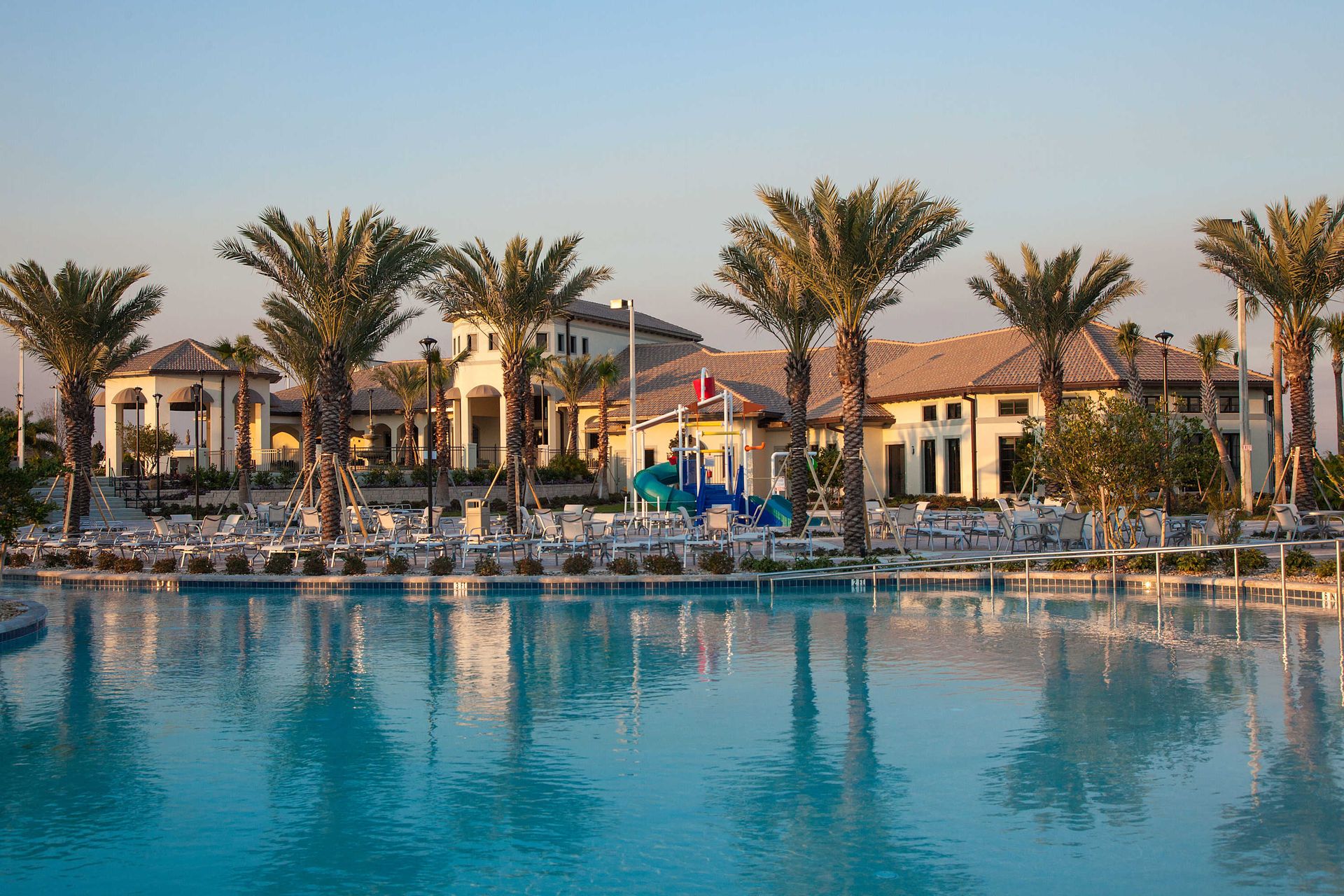 Resort clubhouse, large swimming pool, water play structure, and many palm trees.