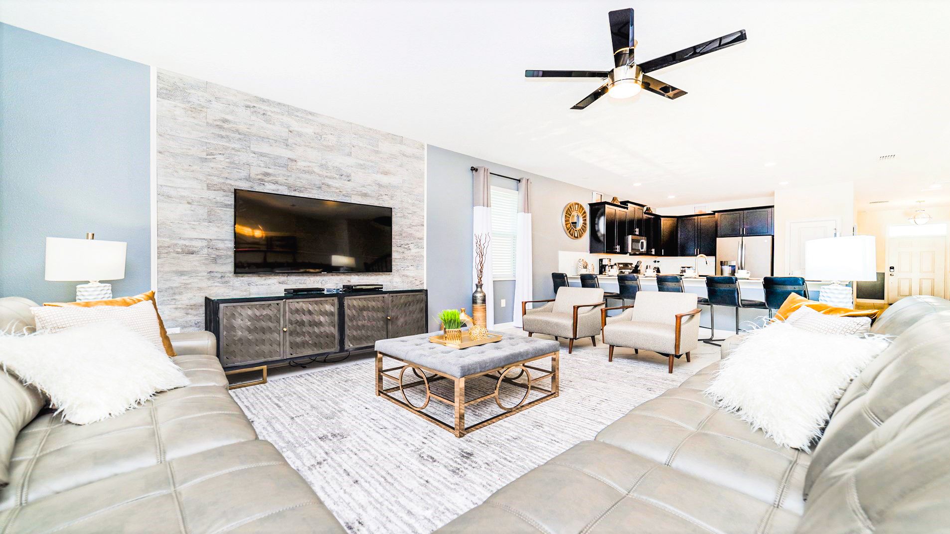 Modern living room with gray accents, a large TV, plush seating, and a sleek kitchen in the background.