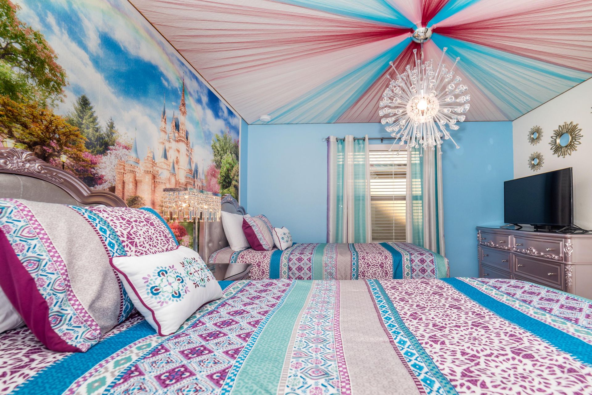 Fairy-tale themed bedroom with vibrant canopy, castle mural, twin beds, and sparkling chandelier.