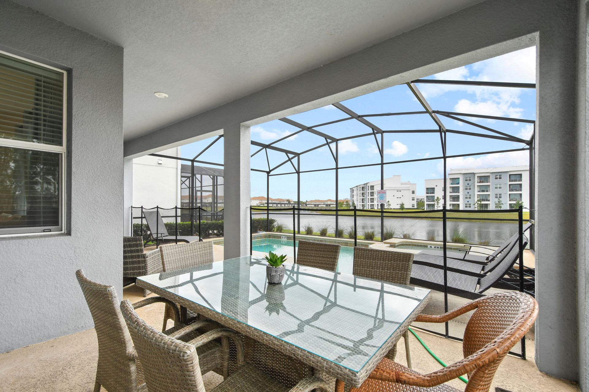 Covered patio dining area with a glass-top wicker table, overlooking a screened pool and spa with tranquil lake views and modern apartment buildings in the distance.