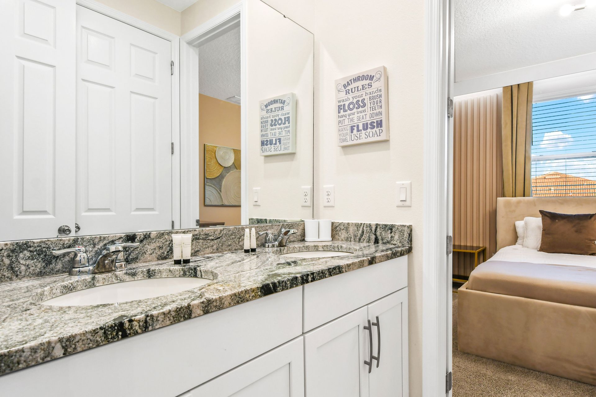 Bright bathroom with a granite double-sink vanity and a view into a cozy, elegant bedroom.