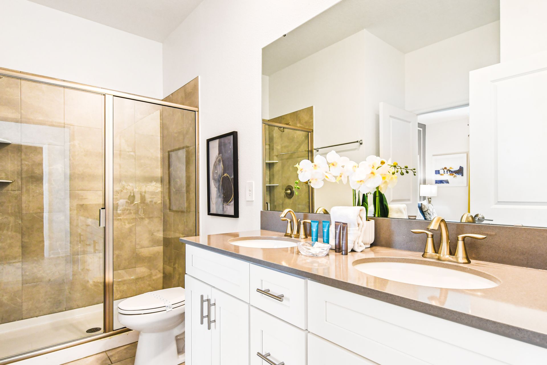 Bright bathroom with dual sinks, gold fixtures, a tiled shower, and chic decor accents.